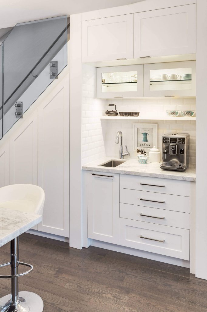 Designer Lisa Robazza utilized the space under the stairs in this Canadian kitchen to add a coffee bar and sink. Space features a marble countertop, a subway tile backsplash, and a floating shelf.