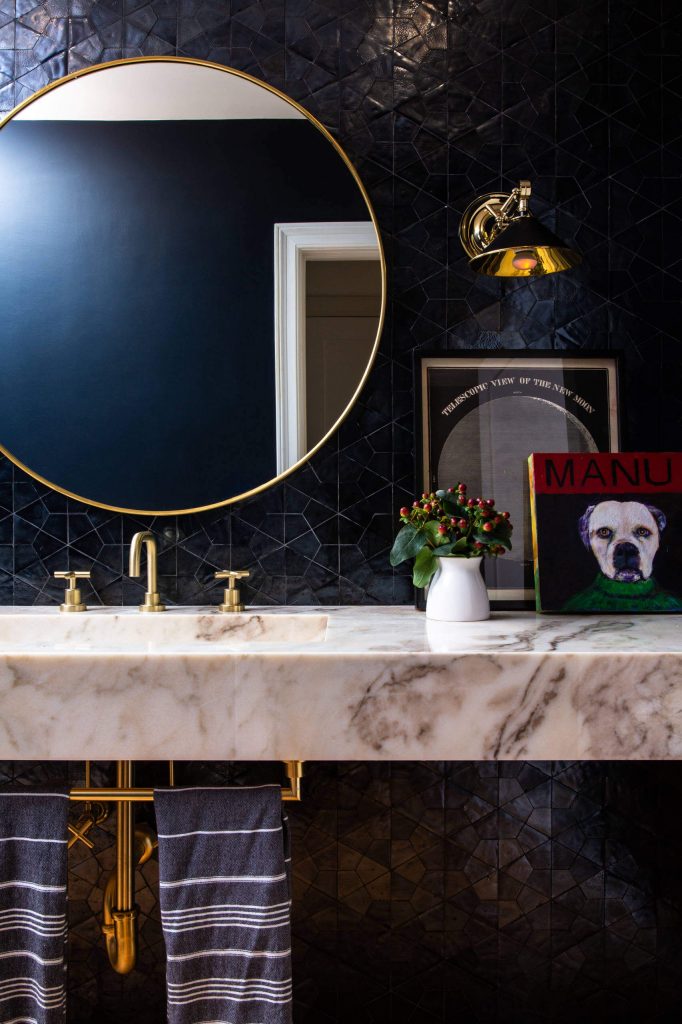 This dark and moody Los Angeles powder room designed by JAC Interiors has some glam rock-star appeal with its sleek black tiled walls, floating marble vanity, and funky accessories.