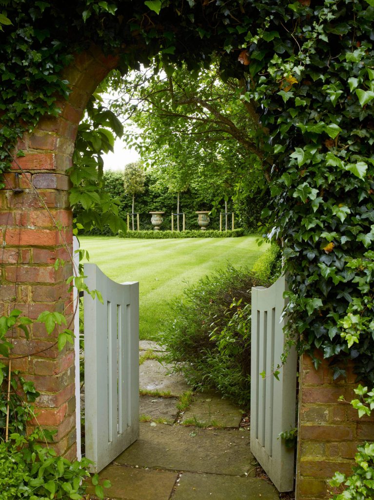 A brick arbor and swinging white garden gates lead to a grassy garden in Sussex, England.